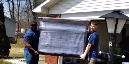 Two men moving wrapped furniture outdoors.